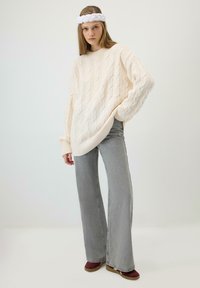 Noor naine pika juustega kannab valget soonikkoes kampsunit, halli laiade sääreosadega teksaseid, burgundiapunaseid kingi ja valget peapaela, ta seisab tavalise tausta ees.