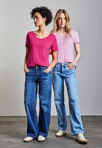 Dos modelos llevan una camiseta rosa y otra lila clara, combinadas con vaqueros anchos de color azul. Ambas tienen las manos en los bolsillos, mostrando un estilo casual.