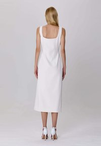 Femme de dos, portant une robe midi sans manches blanche et des chaussures à talons blancs sur un fond uni.