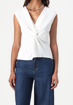 Biała bezrękawnikowa bluzka z wiązanym detalem w talii, zestawiona z niebieskimi szerokimi spodniami jeansowymi, prezentująca gładką, miękką fakturę.