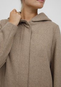 Hnědý kabát ze směsi vlny s kapucí, s jemným texturovaným povrchem a předním zapínáním. Viditelné stehy podél švů.