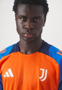 Camisola laranja com detalhes em azul e marinho, apresentando um logótipo branco no peito e detalhes em riscas brancas nas mangas. Tecido sintético e suave.