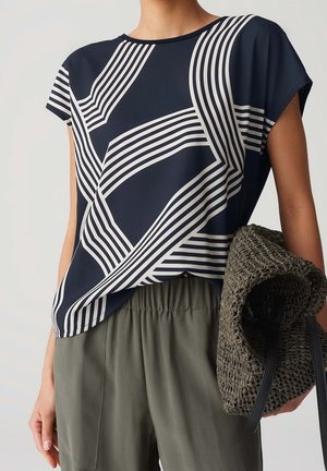 Navy blue short-sleeve blouse with white geometric lines, made of a smooth fabric. Paired with olive green elastic-waist trousers and a woven bag.