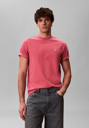 Junger Mann steht, trägt ein pinkes T-Shirt mit hochgekrempelten Ärmeln und verstecktem Logo sowie dunkelgraue Jeans, eine Hand in der Tasche, vor einfarbigem Hintergrund.