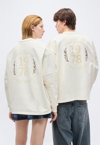 Δύο άτομα κοιτούν πλάγια φορώντας λευκές φούτερ με το κείμενο "CARLO COLUCCI 1978" και σχέδια δάφνης στην πλάτη, σε συνδυασμό με τζιν παντελόνια.