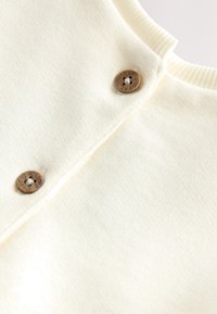 Cremefarbenes Strickgewebe mit glatter Textur, versehen mit zwei runden Holzknöpfen und einem gerippten Kragen.