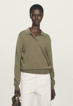 Vrouw in een licht olijfgroene lange mouwen polo trui en witte broek, houdt bruine sandaaltjes met banden vast tegen een effen achtergrond.