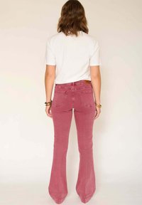 Jean flare ajusté dans une couleur rose poussiéreux douce, fabriqué en denim, avec des poches arrière et une étiquette de marque marron au-dessus de la taille.