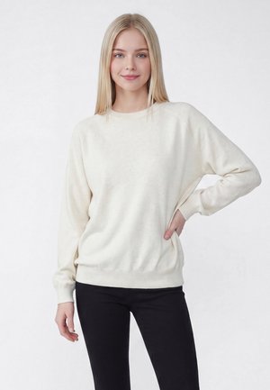 Ung kvinde med langt blondt hår iført en hvid sweater og sorte bukser står med den ene hånd i siden mod en ensfarvet baggrund.