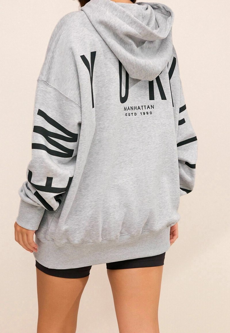 Sudadera gris con capucha, que presenta el texto negro "NUEVA YORK" y "MANHATTAN ESTD 1990" en la parte posterior, y mangas largas y holgadas.