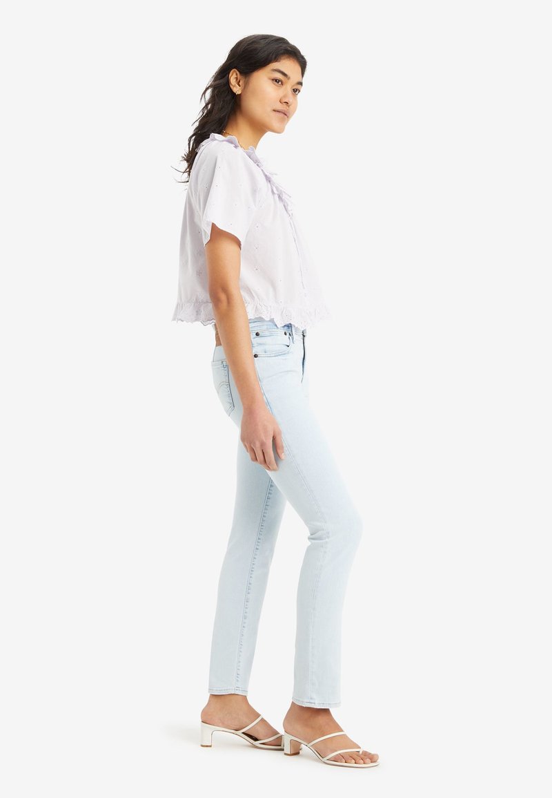 Junge Frau mit langen Haaren trägt eine weiße kurzärmelige Bluse, hellblaue Jeans und weiße flache Sandalen. Sie steht seitlich vor einem weißen Hintergrund.