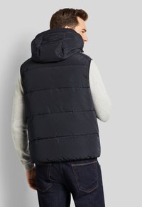 Dunkelblauer Steppwesten mit Kapuze, ausgestattet mit gesteppten Paneelen, einem glatten Außenmaterial und einer taillierten Passform, getragen über einem hellgrauen Oberteil.