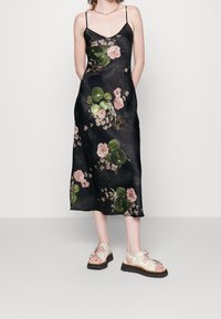 Svart satindress med blommigt tryck i rosa och grönt. Har tunna spaghettiband och en medellång, flödande design. Bärs med ljusa sandaler.