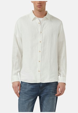 Mężczyzna ubrany w białą koszulę z długim rękawem na białej podkoszulce, niebieskie dżinsy, na jednolitym tle.