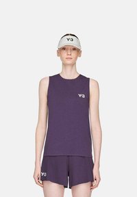 Frau in lila ärmellosem Top und Shorts mit Y-3-Logos sowie einer weißen Y-3-Visiermütze, die vor einem einfarbigen hellen Hintergrund steht.