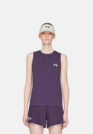 Frau in lila ärmellosem Top und Shorts mit Y-3-Logos sowie einer weißen Y-3-Visiermütze, die vor einem einfarbigen hellen Hintergrund steht.