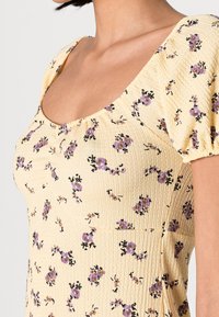 Haut floral jaune avec un tissu texturé, décolleté carré et manches bouffantes ornées de motifs de fleurs violettes et noires.