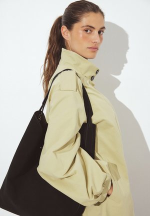 Gaiši dzeltena jaka ar augstu apkakli un melniem pogām, kombinēta ar melnu rokas somu. Abiem priekšmetiem ir gluda tekstūra un izteiktas formas.