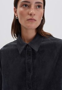 Jonge vrouw met lichte sproeten, die zilveren creolen draagt en een donker corduroy overhemd met knopen aan heeft, en iets opzij kijkt.
