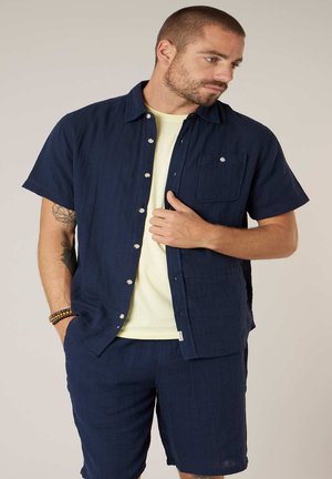 Man met kort haar en baard, gekleed in een marineblauw overhemd met korte mouwen, bijpassende korte broek, lichtgeel T-shirt en armbanden, kijkt naar rechts.