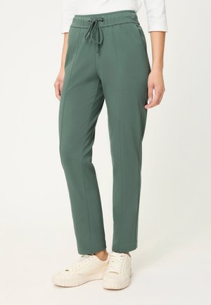 Frau trägt grüne Hosen mit Kordelzug und weiße Turnschuhe, dargestellt von der Taille bis zu den Füßen vor einem einfarbigen Hintergrund.