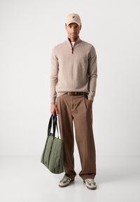 Beige zip-neck sweater with red accent, brown trousers, green quilted tote bag, and beige baseball cap. Sneakers with green and white accents.