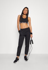 Canotta sportiva nera con fascia con logo, abbinata a pantaloni della tuta neri. Gli accessori includono fasce da polso e una corda per saltare. Le scarpe da ginnastica nere completano il look.