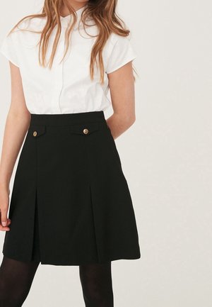 Jeune personne portant un chemisier blanc à manches courtes rentré dans une jupe plissée noire avec des boutons décoratifs dorés et des collants noirs.