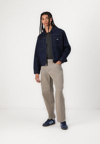 Denim jacket in dark blue with two chest pockets, paired with light beige pants. Model wears navy sneakers with light blue accents.