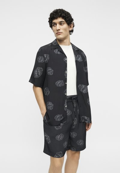 Άνδρας που φοράει μαύρο κοντομάνικο πουκάμισο με κουμπιά και αντίστοιχα σορτς με γκρι λουλουδάτα μοτίβα, στέκεται μπροστά σε λευκό φόντο.