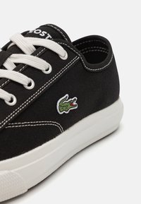 Zapatilla de lona negra con cordones blancos, costuras blancas y suela de goma. Presenta un logo de cocodrilo bordado en verde en el lateral.