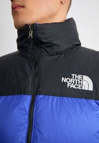Zwarte en blauwe geïsoleerde jas met een hoge kraag, voorzien van een ritssluiting aan de voorkant en een prominent wit logo op de bovenste linkerkant.