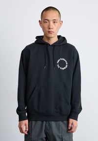 Homme portant un sweat à capuche noir New Balance avec un logo blanc circulaire, se tenant devant un fond gris clair uni, tourné vers l'avant.