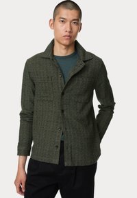Dunkelgrüne, strukturierte Hemdjacke mit Kragen, vorderer Tasche und Knopfverschluss, getragen über einem türkisfarbenen Oberteil und schwarzen Hosen.