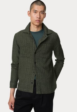 Dark green textured shirt jacket with a collared neckline, front pocket, and button closure, worn over a teal top and black trousers.