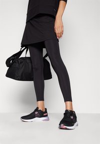 Tenue de sport noire composée d'un haut en couches et de leggings. Les chaussures sont noires avec des accents roses ; un sac de sport noir est tenu dans une main.