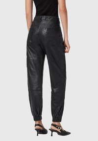 Vrouw in zwarte high-waisted leren joggerbroeken met achterzakken en elastische boorden, gecombineerd met zwarte slingback hakken.