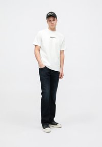 Camisola branca com logotipo preto, jeans de denim azul-escuro e boné preto. Ténis pretos com solas e atacadores brancos, em pé contra um fundo neutro.