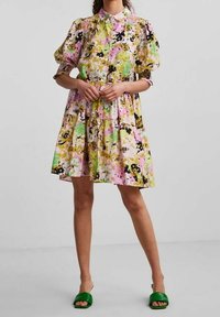 Bloemendress met een kraag, pofmouwen, een gelaagde rok en een mix van roze, geel, groen en zwarte kleuren. Gegdraagd met groene sandalen.