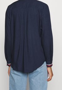 Mörkblå långärmad skjorta med plisserad rygg, kontrasterande randiga manschetter och rundad nederkant. Tillverkad av lättviktigt material för komfort.