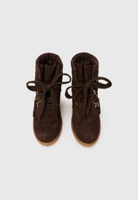 Par de ténis altos de camurça castanho escuro com atacadores grossos e solas de borracha cor de goma sobre um fundo branco.