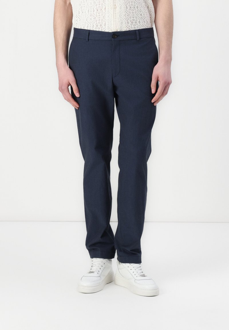 Marineblå bukser laget av glatt stoff, med slim fit, frontknappelukking og side lommer, parret med hvite sneakers.