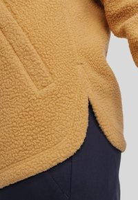 Gelbe Fleecejacke mit strukturierten Oberflächen, mit einem Seitenschlitz und einer Tasche. Trägt sie über dunkelblauen Hosen.