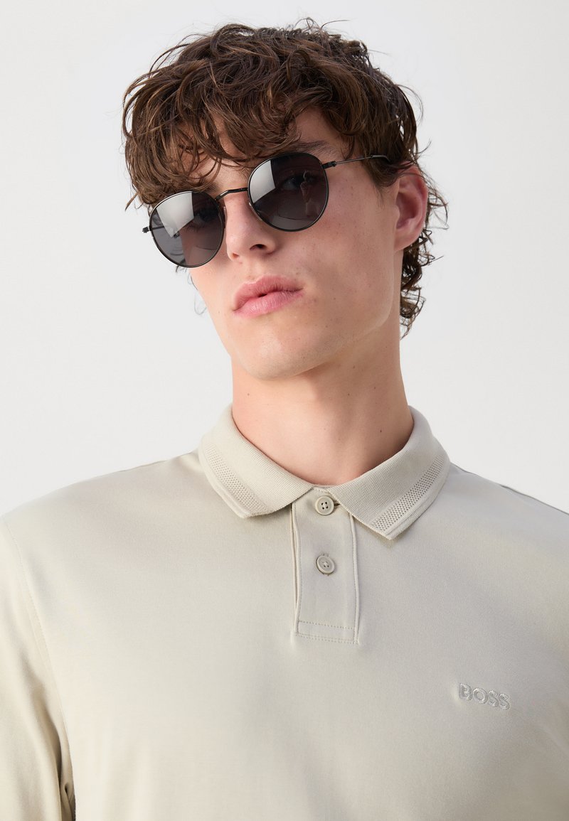 Polo shirt beige pâle avec un col, featuring deux boutons et un logo "BOSS" brodé ; lunettes de soleil rondes noires avec des montures fines.