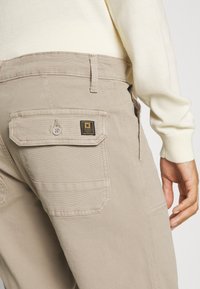 Beige cargo hla�če z zadnjim žepom z gumbom in blagovno znamko. Tkanina ima gladko teksturo in prilega se po meri.