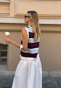 Gilet en tricot rayé bleu marine, rouge et crème sur une jupe blanche évasée. Tenant un cornet de glace. Bracelet en argent au poignet.
