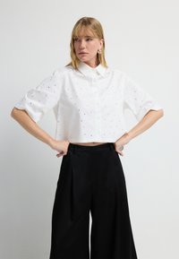 Camisa blanca recortada de botones con mangas cortas, que presenta pequeños puntos decorativos negros. Combinada con pantalones de pierna ancha negros de talle alto.