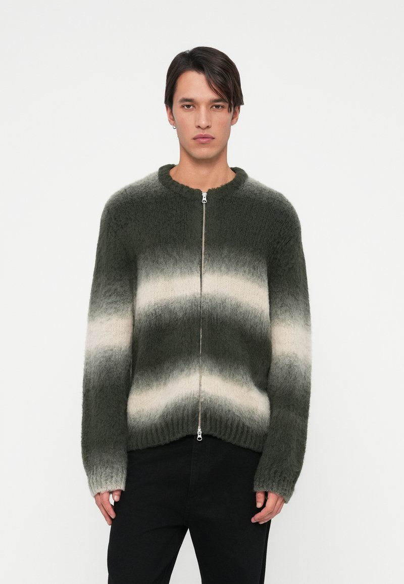 Jovem usando um suéter zíper verde e branco listrado e calças pretas, em pé contra um fundo branco liso.