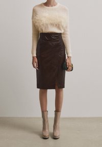 Femme portant un pull crème avec un détail en plume, une jupe crayon en cuir marron foncé avec une fente à l'avant, des bottines beige, et tenant une petite pochette sombre.