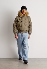 Hombre caminando con una chaqueta de piel con capucha color beige, jeans de pierna ancha azules claros, gorro negro y zapatos negros sobre un fondo blanco liso.